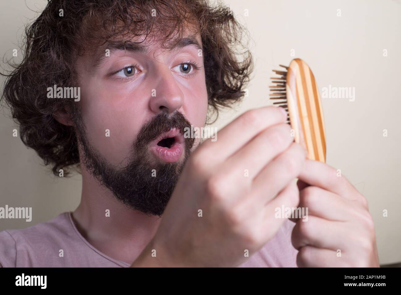 Portrait of a young guy with long dirty hair and problematic scalp. Holding a comb with lost hair, looking at her indignantly. Hair loss at an early a Stock Photo