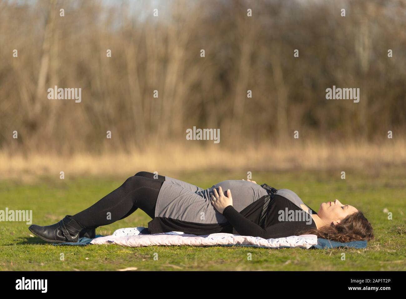 Pregnant woman resting on her back on the grass lawn outdoors in the nature on sunny day, laying on soft blanket on the ground with eyes shut and hold Stock Photo
