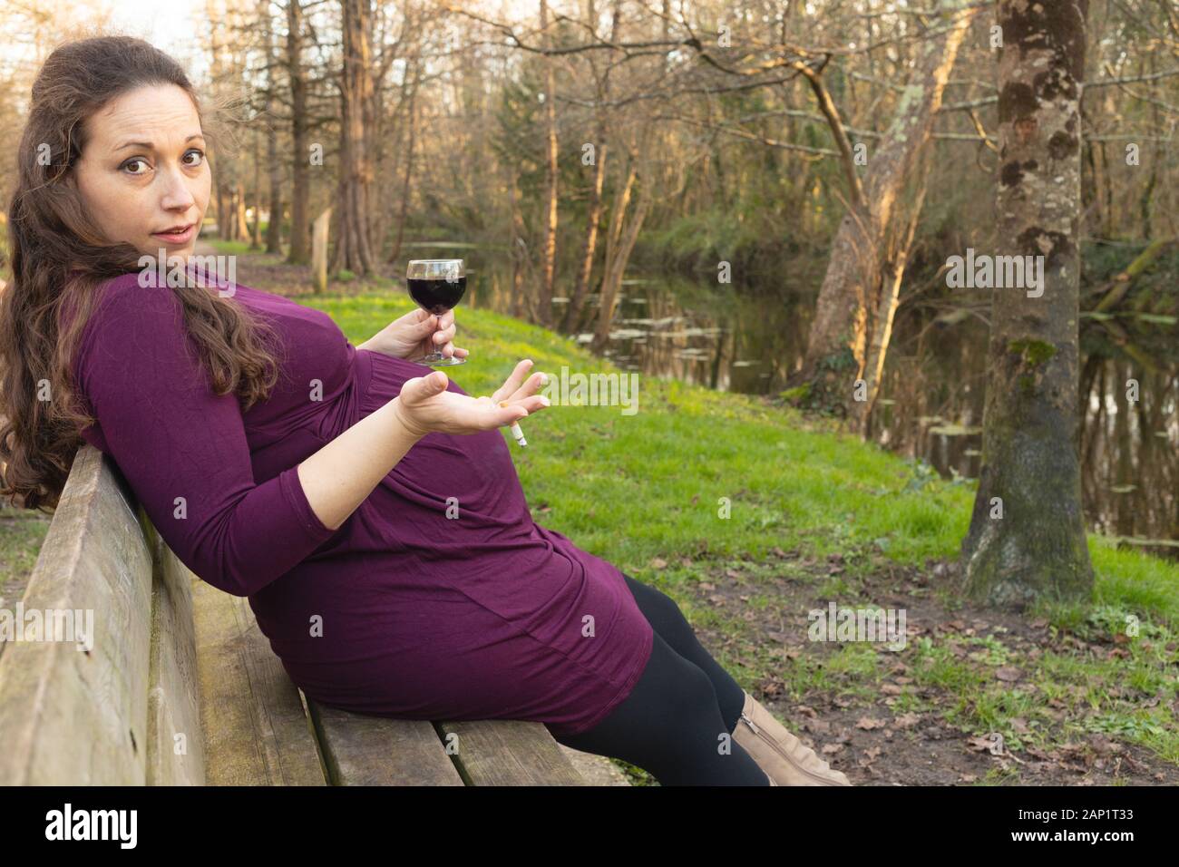 Pregnant woman with alcohol and cigarette in her hands, sitting on the bench in park and looking at camera with questioning look and shrugging gesture Stock Photo