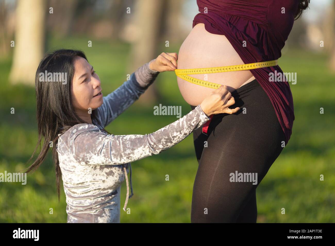 Asian Woman Measuring Pregnant Womans Bare Belly With A Tape Outdoors