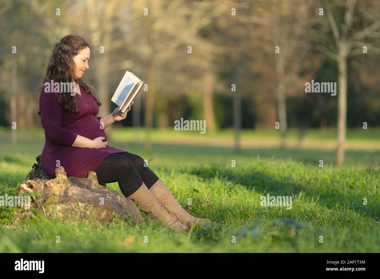 Pregnant woman reading a book, sitting outdoors in the park on sunny day. Full length side portrait with copy space Stock Photo
