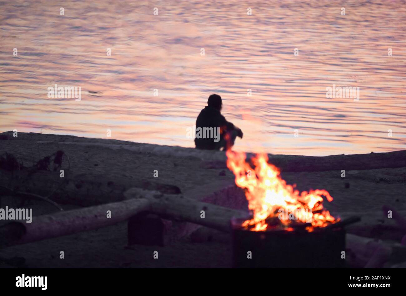 a lone man sits by a campfire on a beach meditating and contemplating his thoughts and staring at the ocean Stock Photo