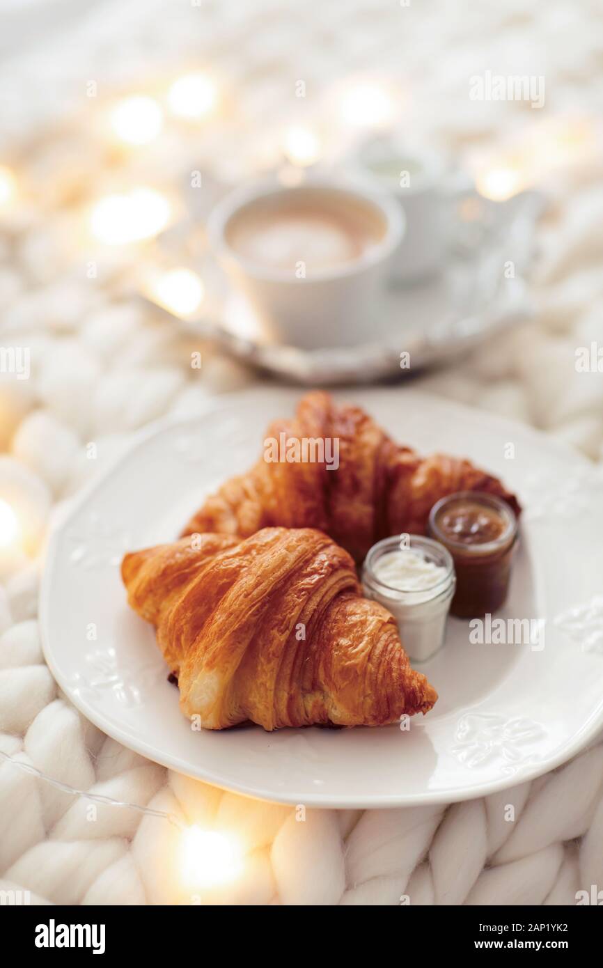 Fresh croissants with jams and americano with milk on knitted white wool blanket and luminous garlands. Cozy winter morning at home. Scandinavian bedroom. Stock Photo
