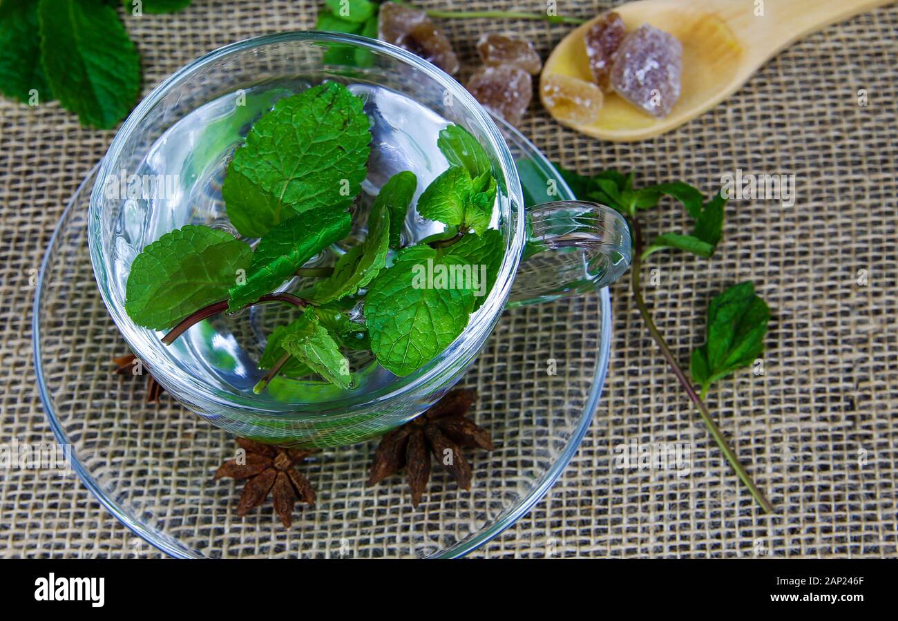 Close up of isolated transparent tea glass cup with fresh mint leaves, wood spoon with brown candy rock sugar and anise stars. jute mat background. Stock Photo