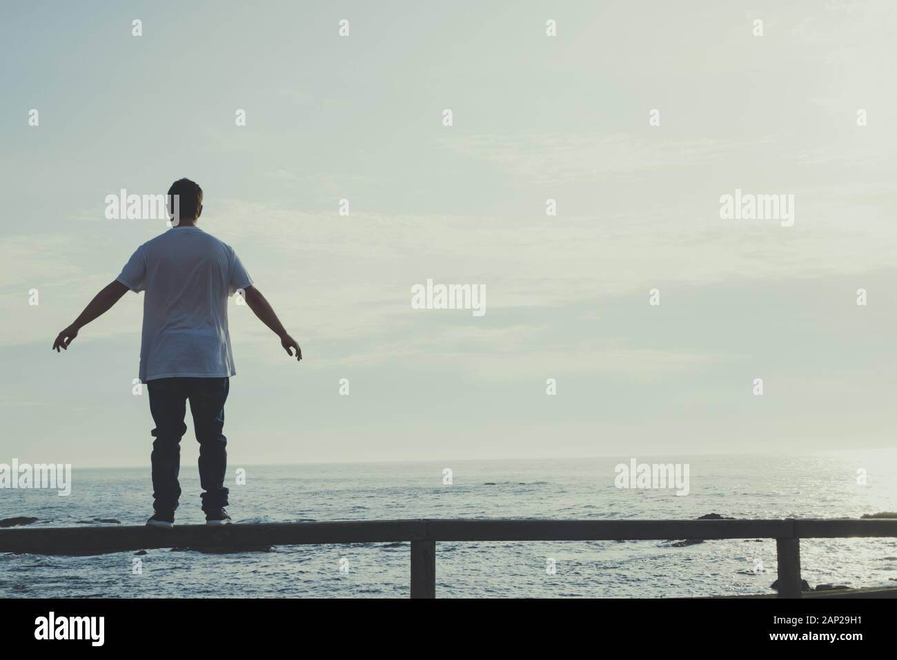 Man with outstretched arms standing on a railing looking at the ocean Stock Photo