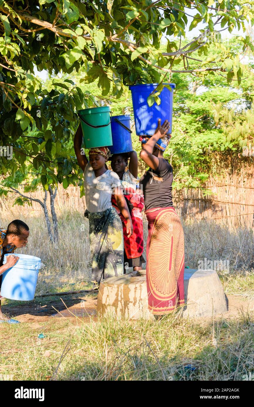 four Malawian women lifting heavy buckets of water onto their heads at the village well ready to carry them back to their homes in the sun Stock Photo