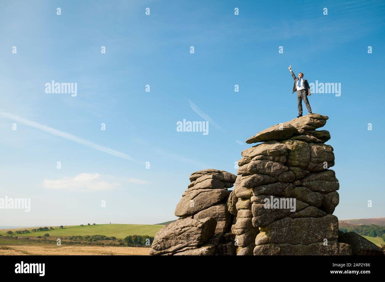Distant businessman standing on the top of a rocky mountain with his finger pointed high in the air Stock Photo