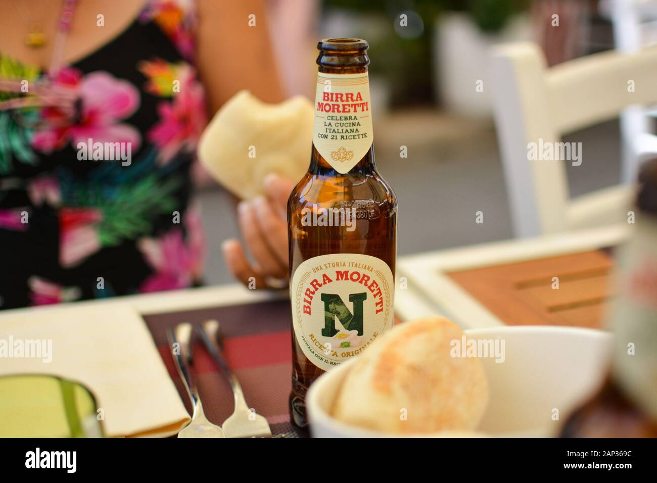 A woman in a flower print top eats a bread roll and enjoys a glass bottle of beer at an outdoor patio in Italy. Stock Photo