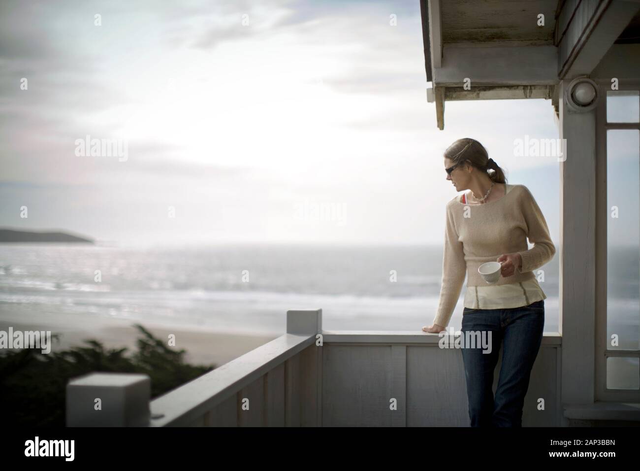 Young woman standing with a tea cup on a balcony by the sea. Stock Photo