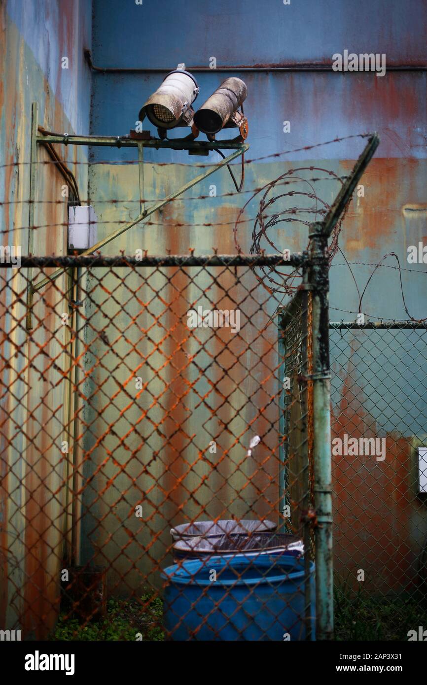 Surveillance cameras atop a chain link fence surrounding a rusty, aging building exterior. Stock Photo