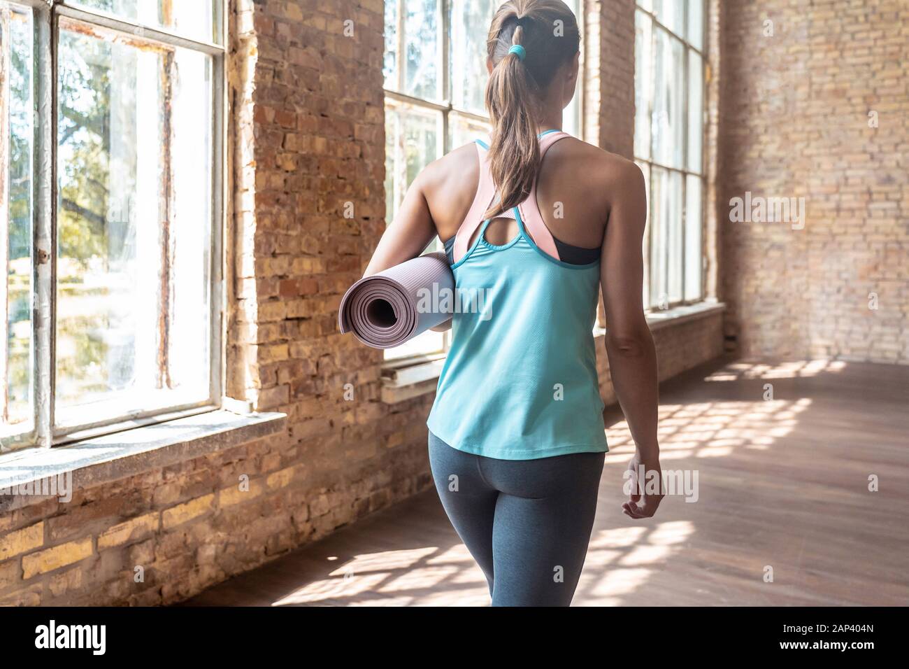 Back view of sporty fit young woman holding rolled yoga mat walking in gym Stock Photo