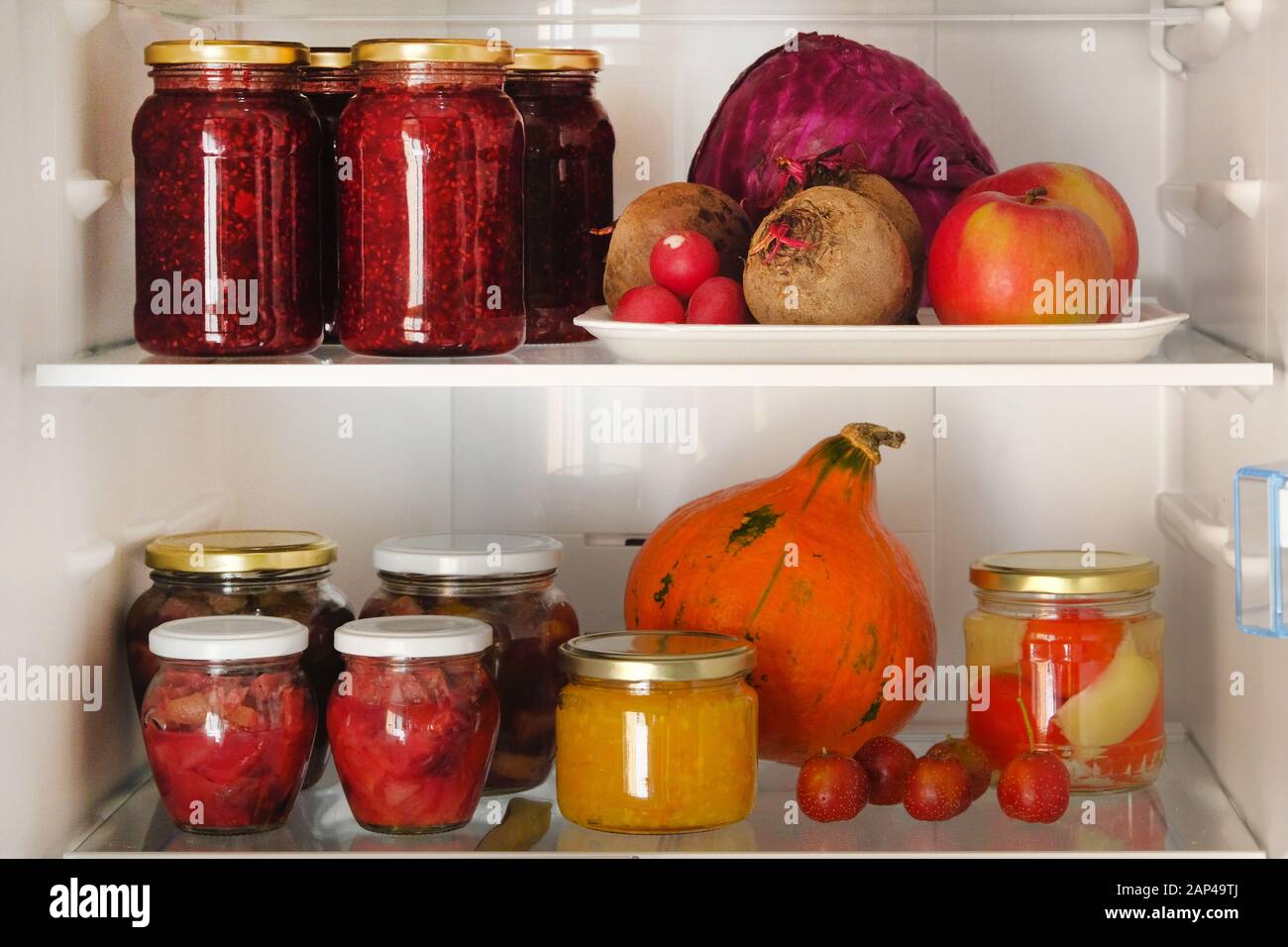 Jars with homemade fruit and berry jams and fresh red, purple and orange vegetables and fruits on shelf in fridge. Fermented healthy vegetarian foods Stock Photo