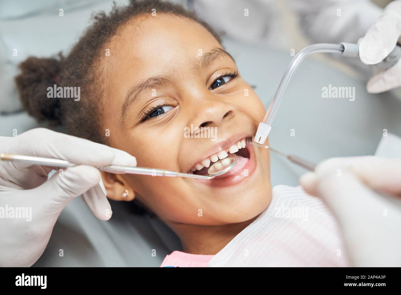Charming little afro american girl sitting in dental chair, smiling and looking at camera during medical treatment at modern clinic. Concept of health care ad pediatrics Stock Photo