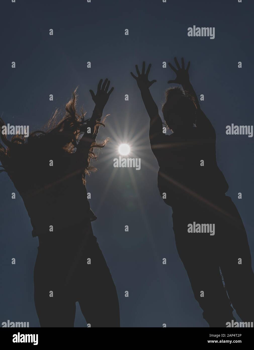 Silhouette of two young women jumping toward the sun with hands up. Dark and low angle image of unrecognizable happy girls. Stock Photo