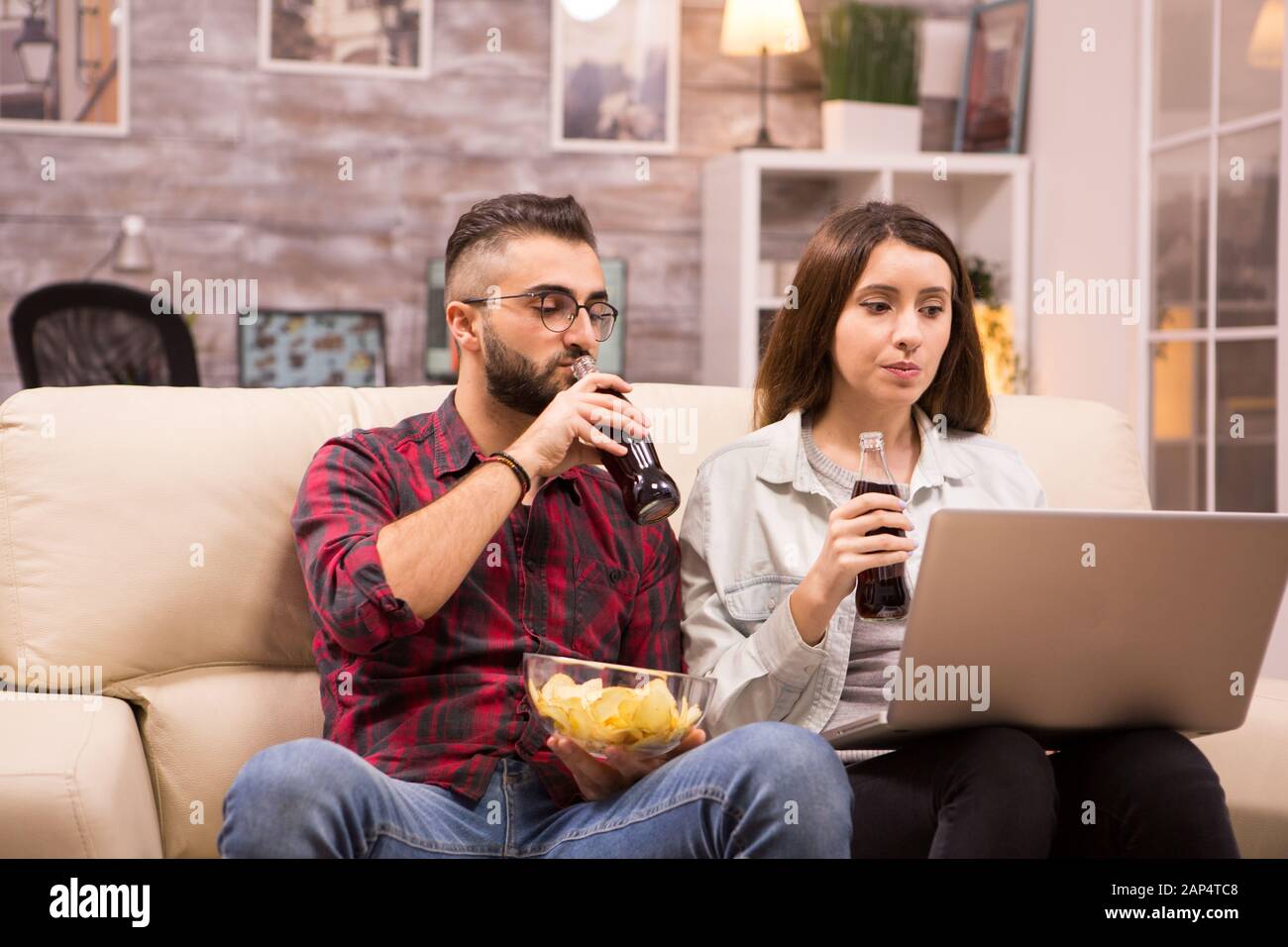 Couple watching a movie sitting comfortable on couch eating chip and drinking soda Stock Photo