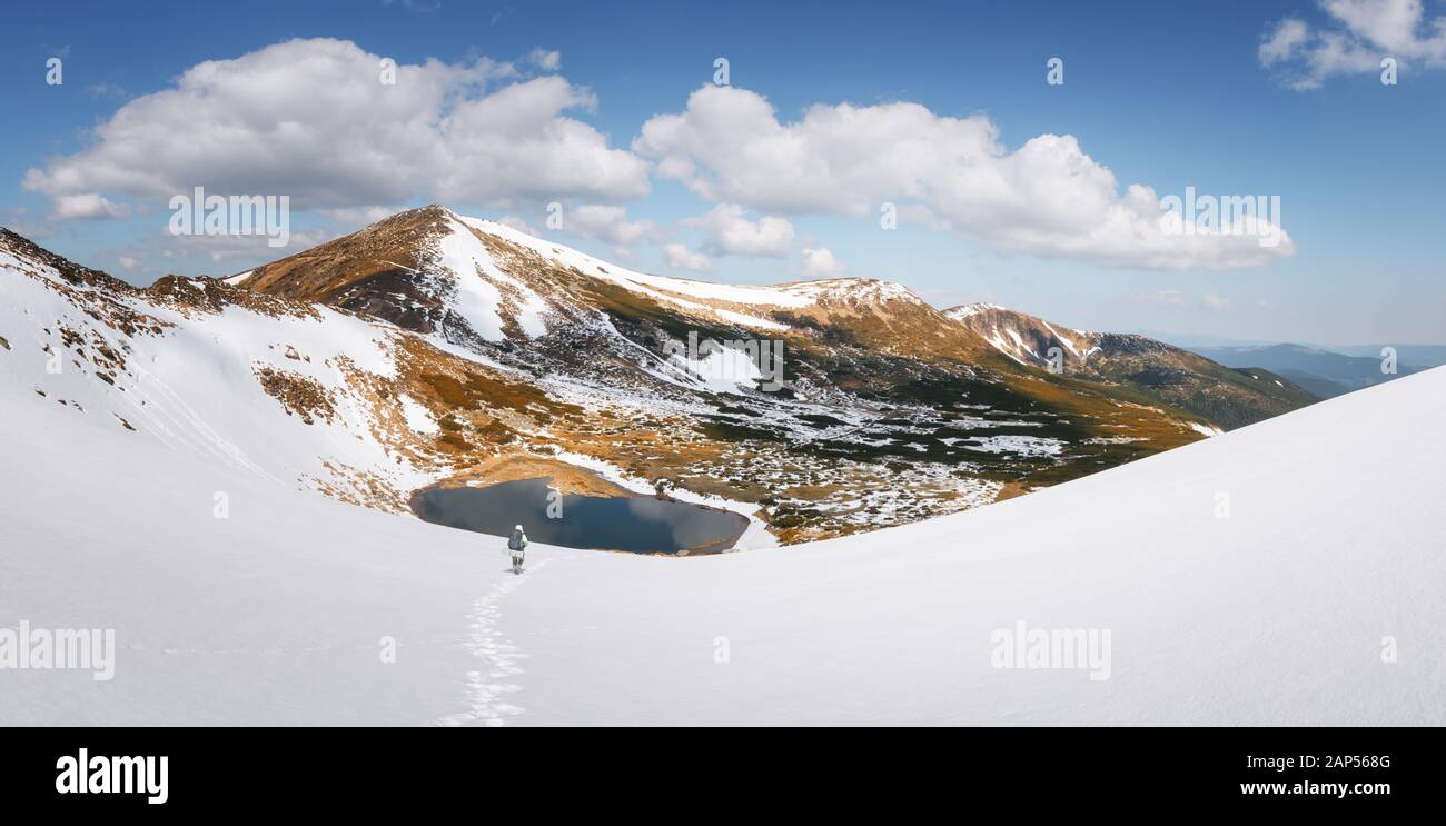 Tourist with a backpack hiking in spring mountains. Frozen mountain lake and snowy hills under a blue cloudy sky. Landscape photography. Panorama Stock Photo