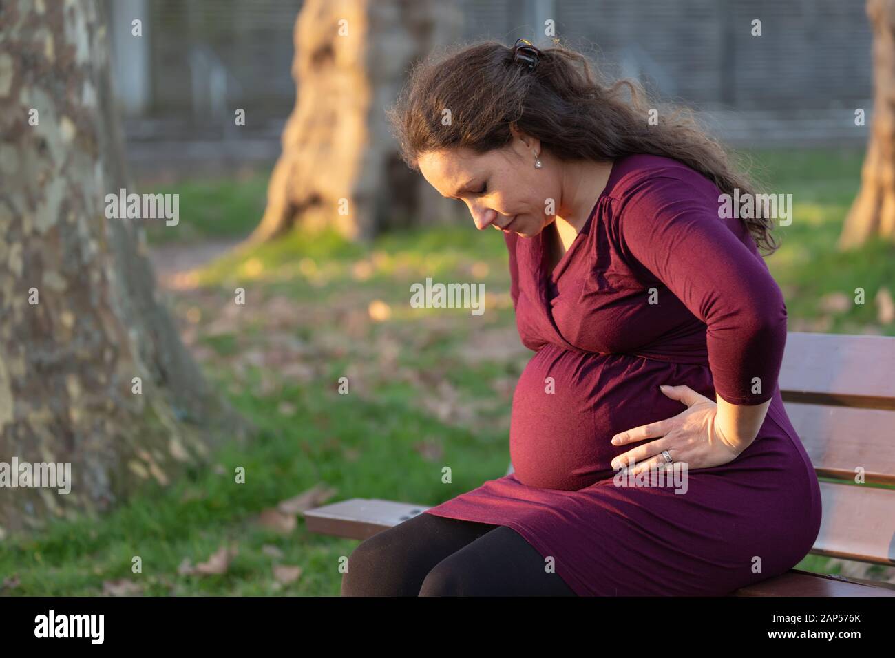 Troubled young pregnant woman on a park bench clasping her swollen belly and leaning forwards with a grimace and eyes closed trying to relax in warm e Stock Photo