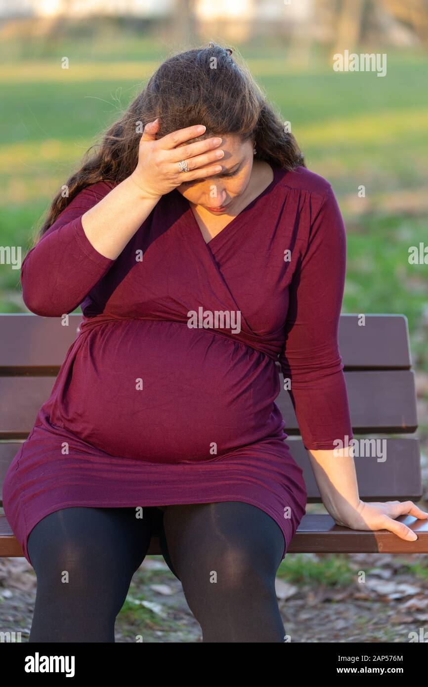 Young pregnant woman suffering from morning sickness holding her hand to her forehead as she relaxes outdoors on a park bench in close up Stock Photo