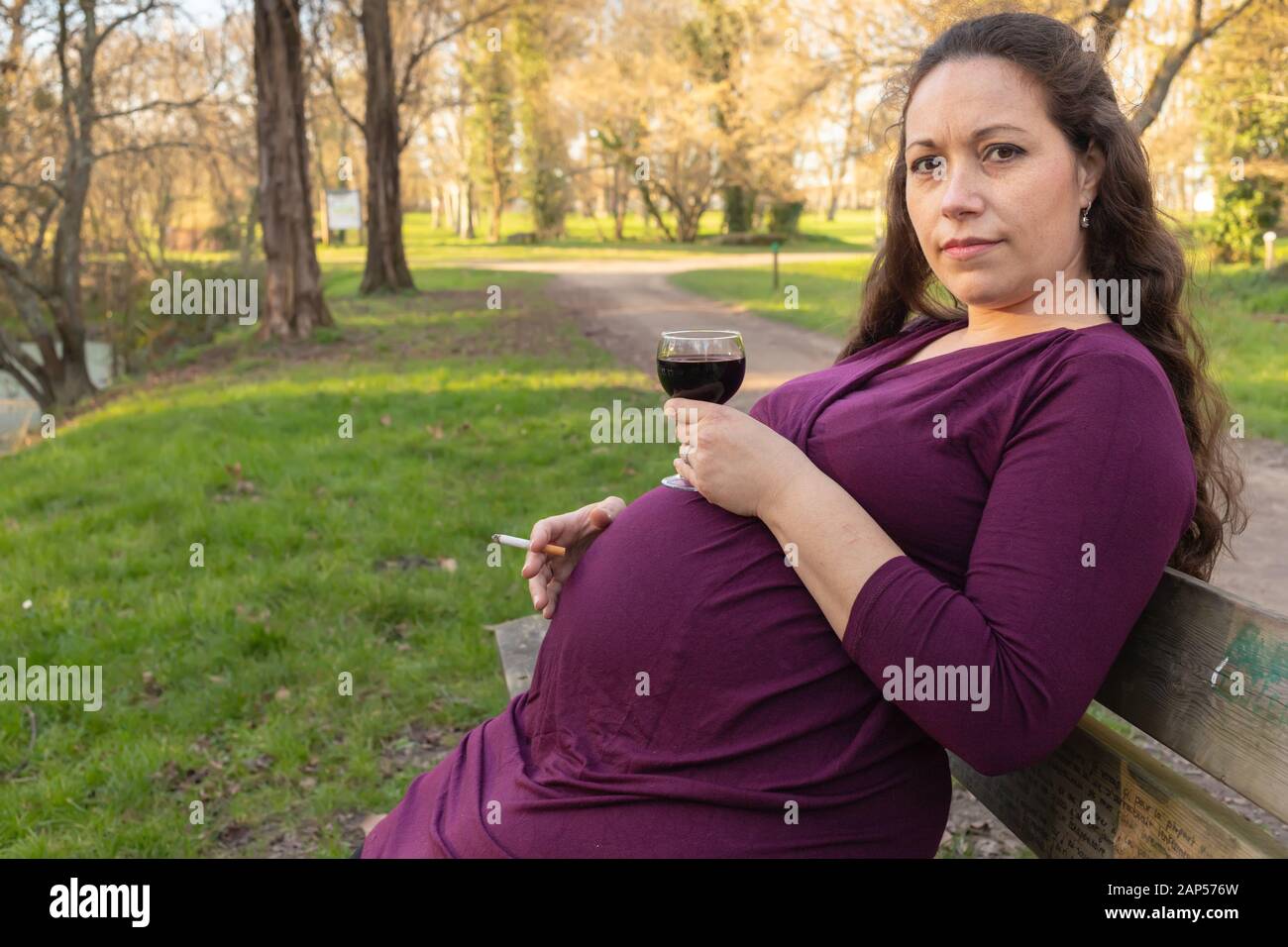 Pregnant woman with alcohol and cigarette in her hands, sitting on the bench in park and looking at camera with questioning look . Side portrait with Stock Photo