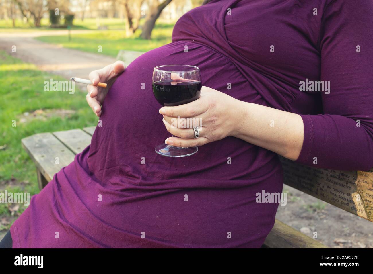 Pregnant woman sitting on a bench, drinking red wine and smoking cigarette in a park. Stock Photo