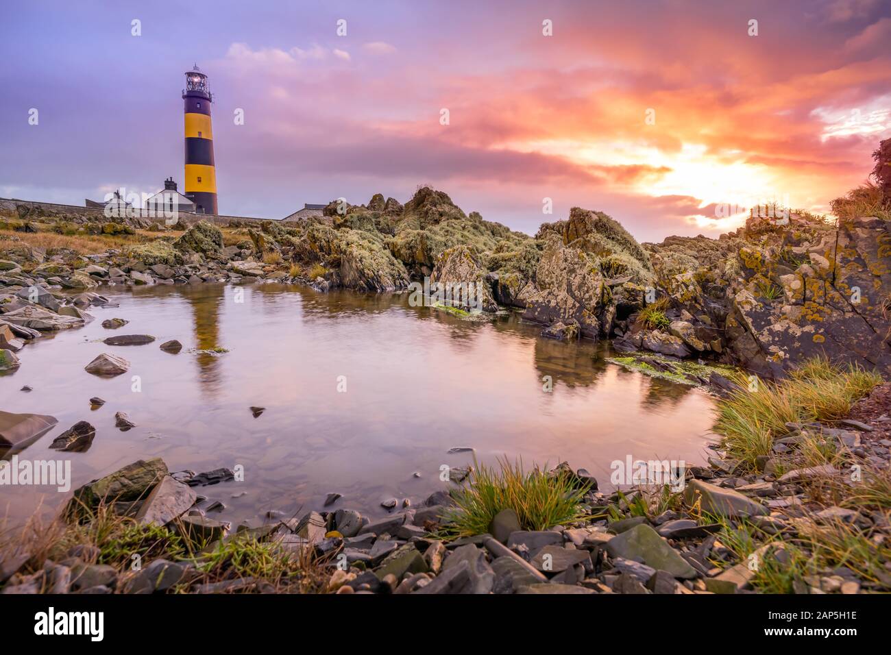 Amazing sunrise at St. Johns Point Lighthouse in County Down, Northern Ireland. Rocks reflected on small pond on coastline. Stock Photo