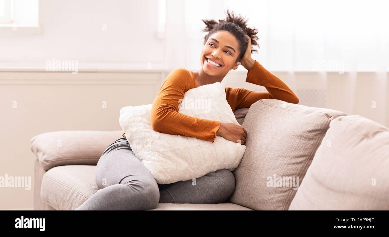 African American Woman Sitting On Sofa Embracing Pillow Indoor, Panorama Stock Photo