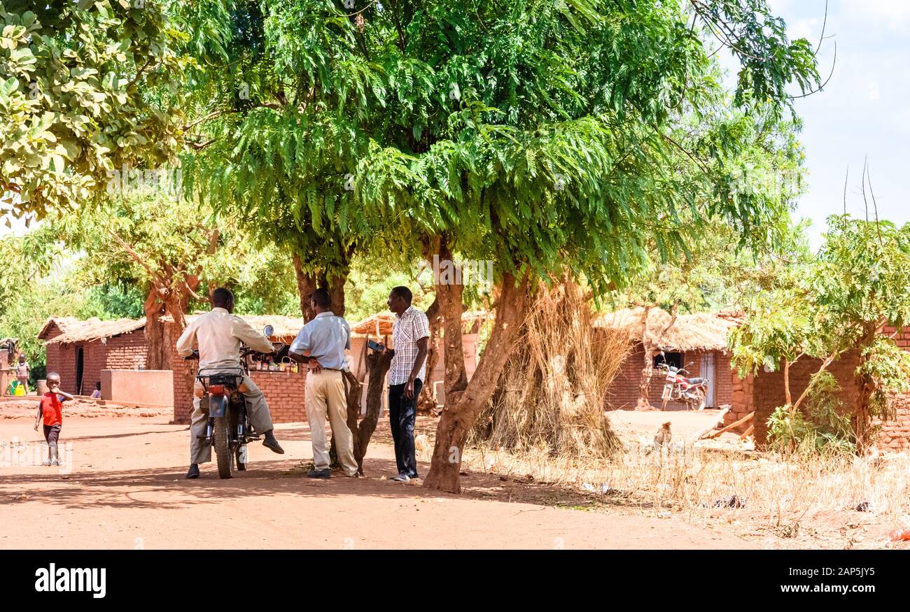 Malawian men stand talking under a large tree for shelter from the hot sun in the centre of a township of Lilongwe one astride a motorcycle Stock Photo