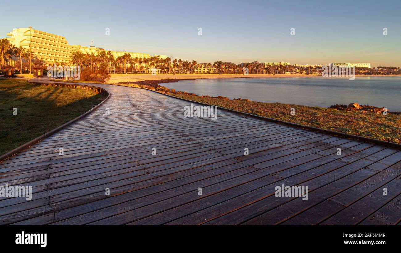 Wooden Boardwalk at sunrise leading to holiday resort lit by morning sun, beach and bay with hotels, restaurants, cafes and palm trees, Sa Coma, Mallo Stock Photo