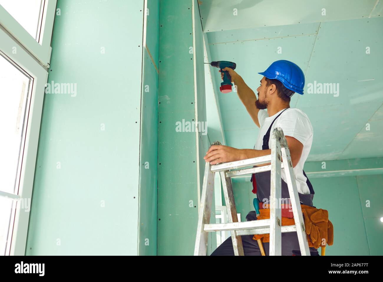 A builder standing on a ladder installs drywall at a construction site ...