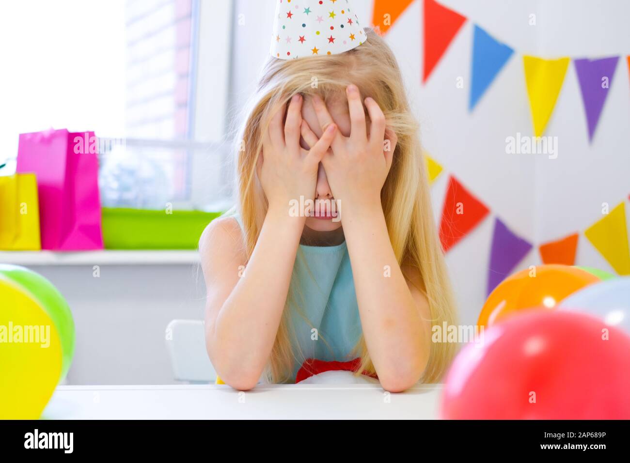 Unhappy blonde caucasian girl is crying near birthday rainbow cake
