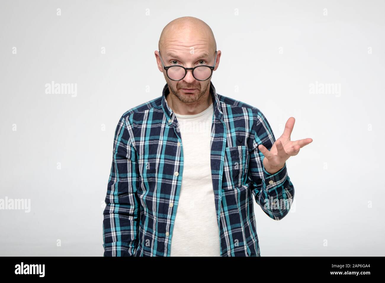 Bald, unshaven man looks inquiringly and calmly into the camera over glasses and makes a hand gesture. Stock Photo