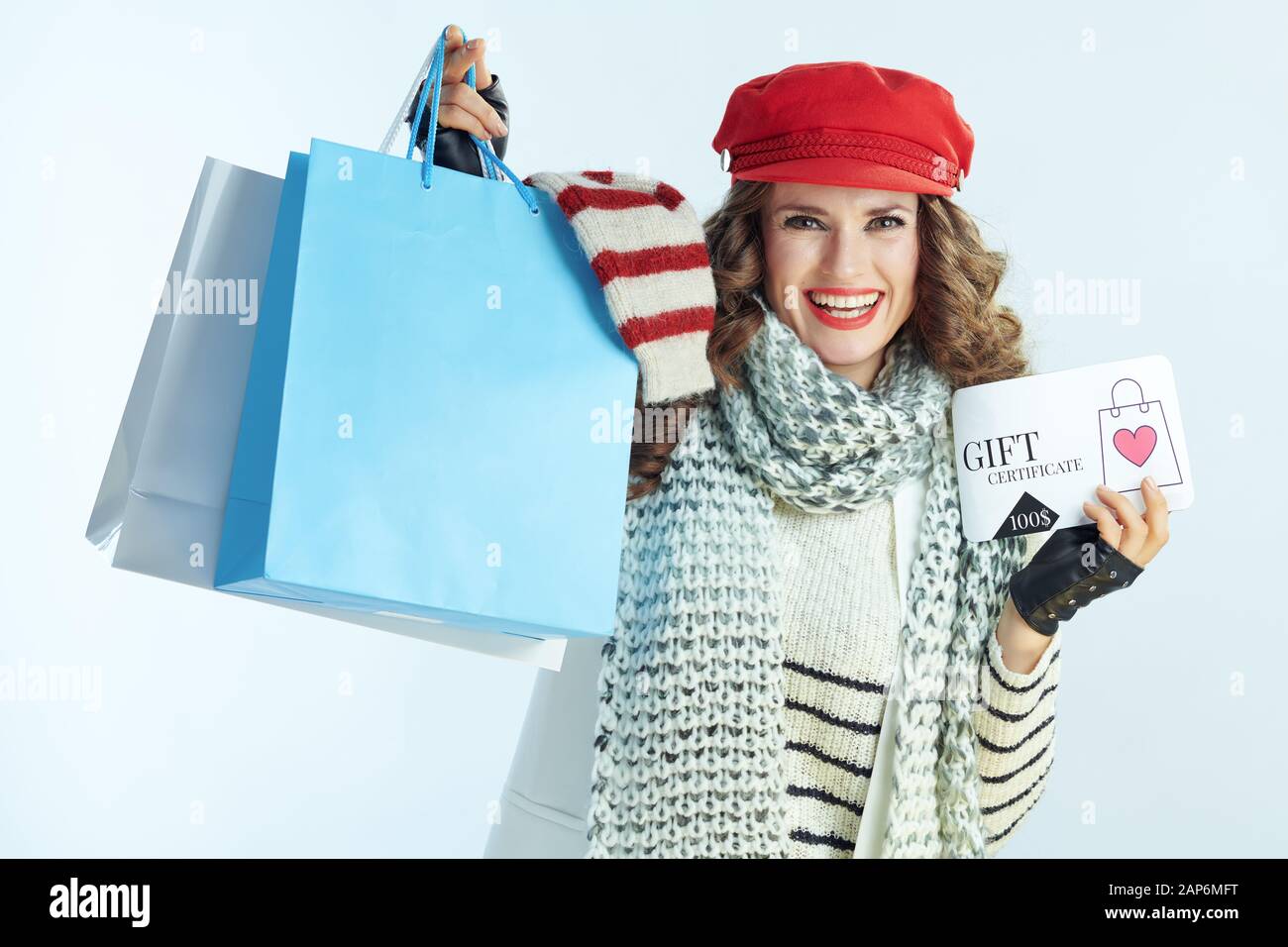 smiling young female with long brunette hair in sweater, scarf and red hat showing shopping bags with sweaters and discount coupon against winter ligh Stock Photo