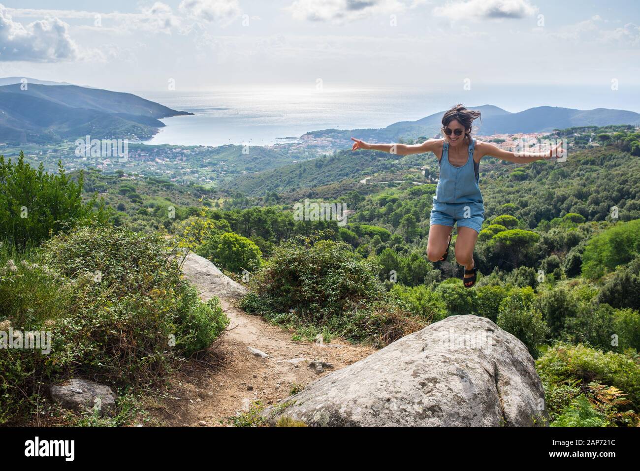 Young woman wearing denim summer dungaree shorts and sandals jumping on rock overseeing Mediterranean sea bay in Tuscany, Italy Stock Photo