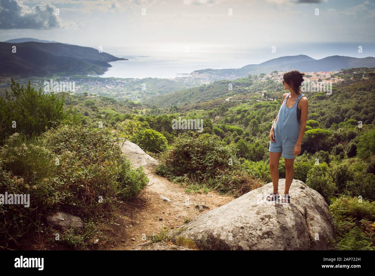 Young woman on holiday wearing denim summer dungaree shorts and sandals admiring panorama from hill overseeing bay in Tuscany, Italy Stock Photo