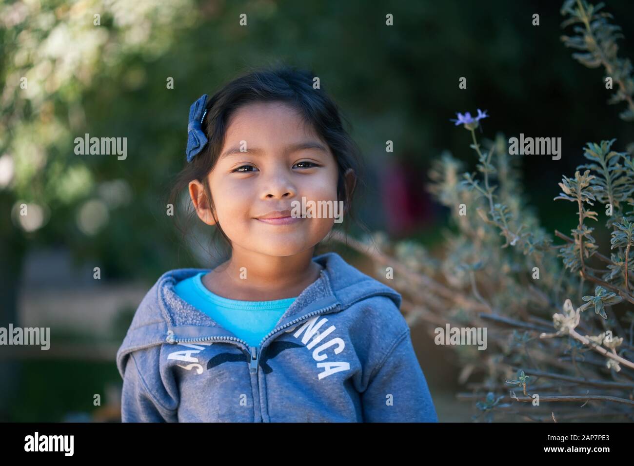 Confident little girl wearing a sweater, standing in front of trees and green plants with a sincere smile on her face. Stock Photo
