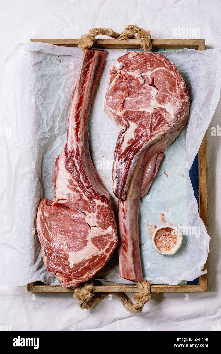 Raw uncooked black angus beef tomahawk steaks on bones served with pink salt on wooden tray with baking paper over white cloth as background. Top view Stock Photo