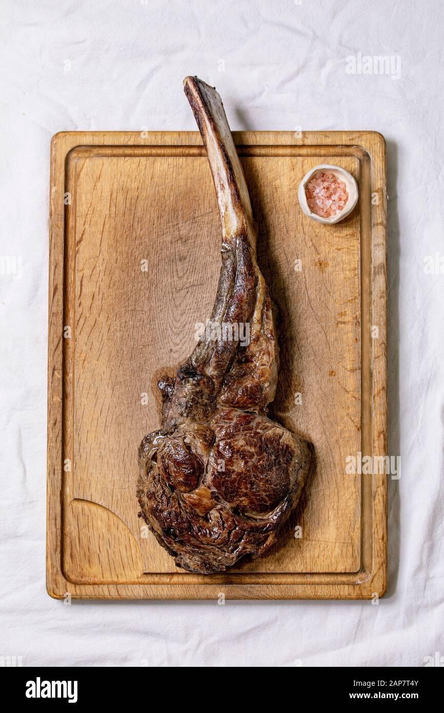 Grilled black angus beef tomahawk steak on bone served with pink salt on wooden cutting board over white cloth as background. Top view, space. Stock Photo