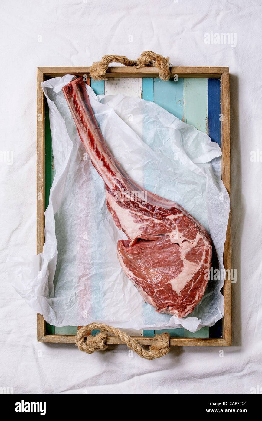 Raw uncooked black angus beef tomahawk steak on bones served on wooden tray with baking paper over white cloth as background. Top view, space. Stock Photo