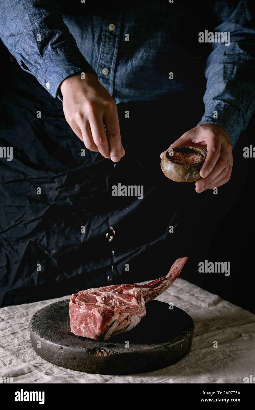 Man's hands sprinkled with salt raw uncooked black angus beef tomahawk steak on bones on linen table cloth. Rustic style Stock Photo