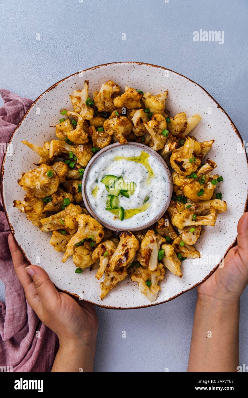 Hands holding roasted spicy cauliflower wings in a ceramic bowl served with a small bowl of yogurt cucumber dip Stock Photo