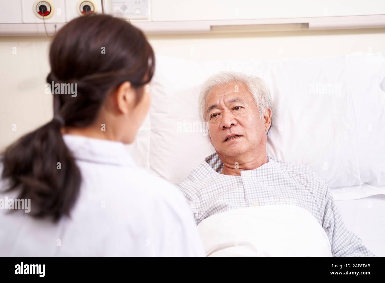 asian old man lying in bed talking to young female doctor in hospital ward Stock Photo