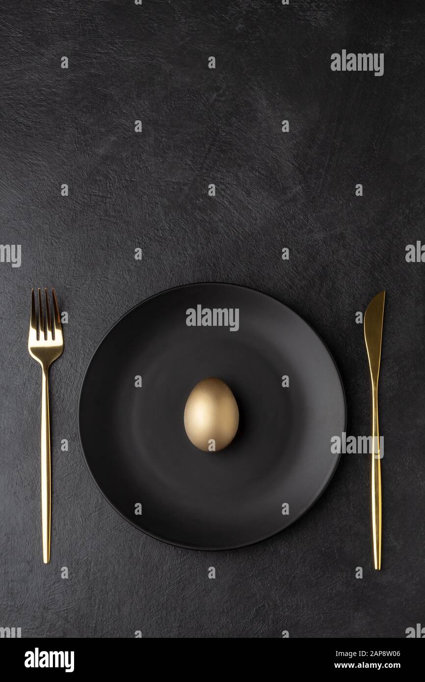 Golden egg on a black plate with a knife and fork. Flat lay with place for text, vertical orientation. Stock Photo