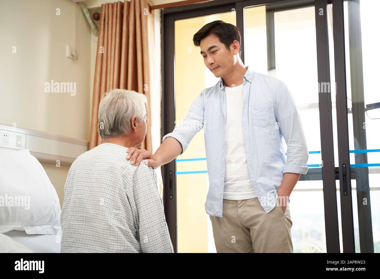 young asian adult son visiting talking to senior father in nursing home or hospital ward Stock Photo
