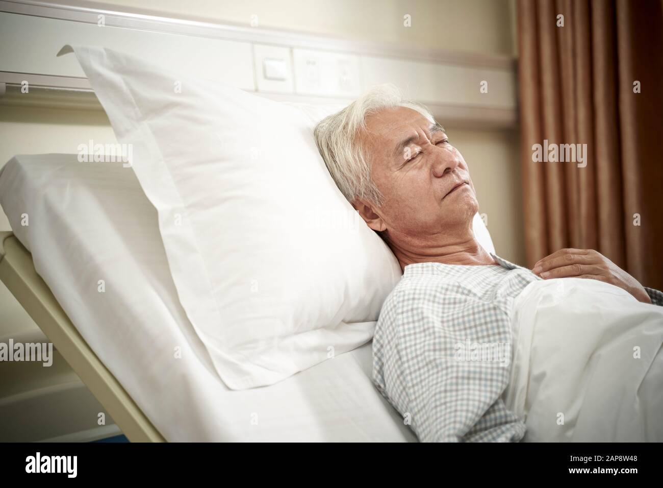 asian elderly male patient lying in bed sleeping in hospital ward or assisted living facility Stock Photo