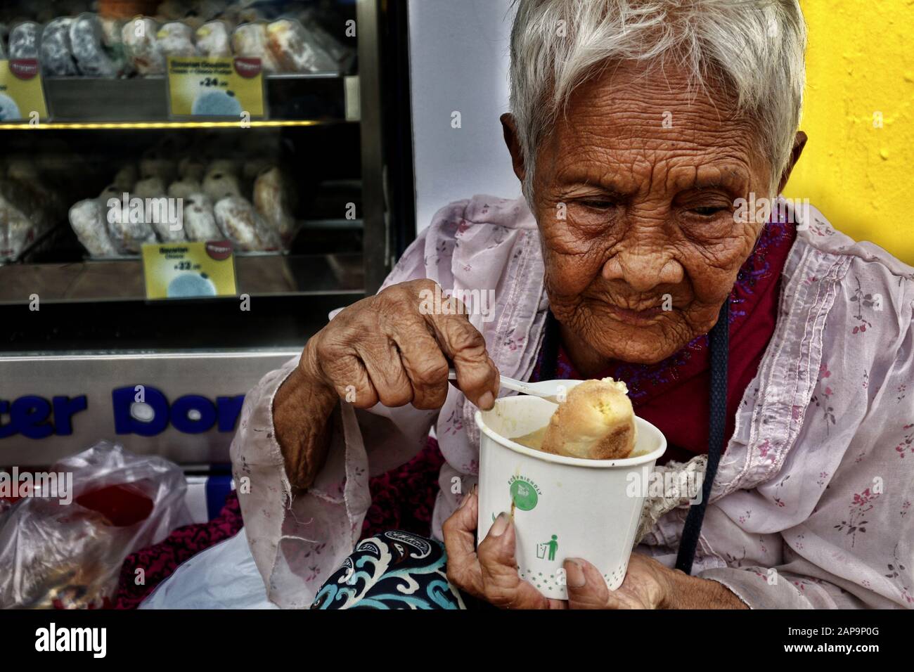 Antipolo City, Philippines - January 18, 2020: Old Filipino woman with wrinkled skin eat a bowl of porridge while sitting on a sidewalk.. Stock Photo