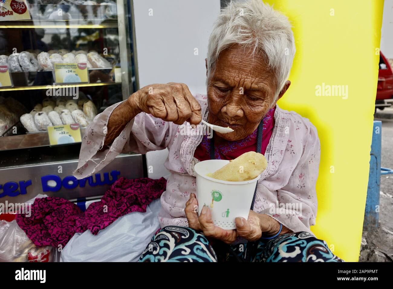 Antipolo City, Philippines - January 18, 2020: Old Filipino woman with wrinkled skin eat a bowl of porridge while sitting on a sidewalk. Stock Photo