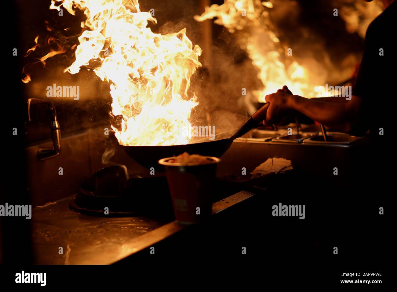 The cook in the kitchen holds a wok with an open flame in his hands