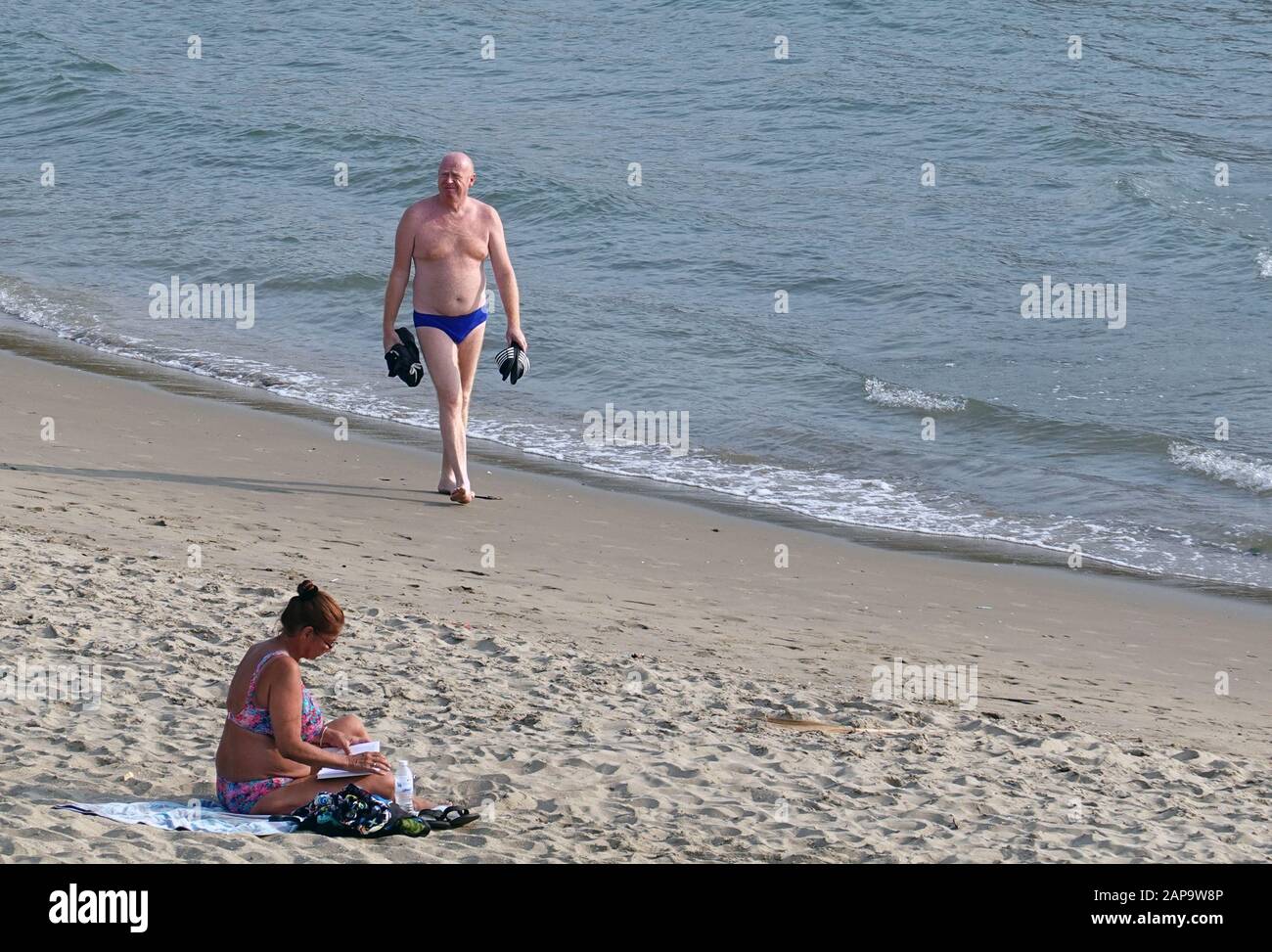 Pattaya, Thailand - December 23, 2019: Bald older man walking on the beach. Woman sits and reading a book. Stock Photo