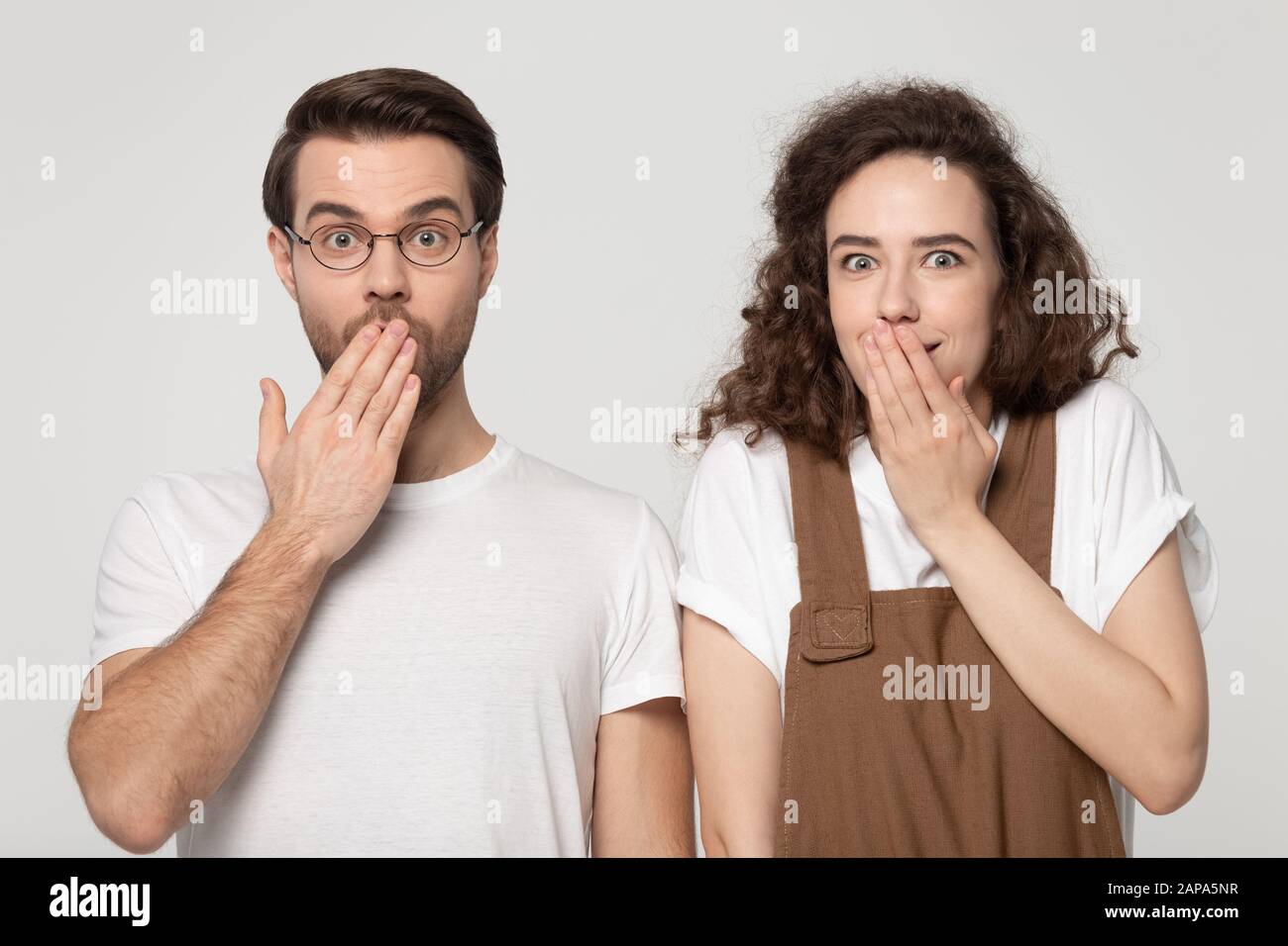 Surprised millennial joyful shy couple covering mouths with hands. Stock Photo