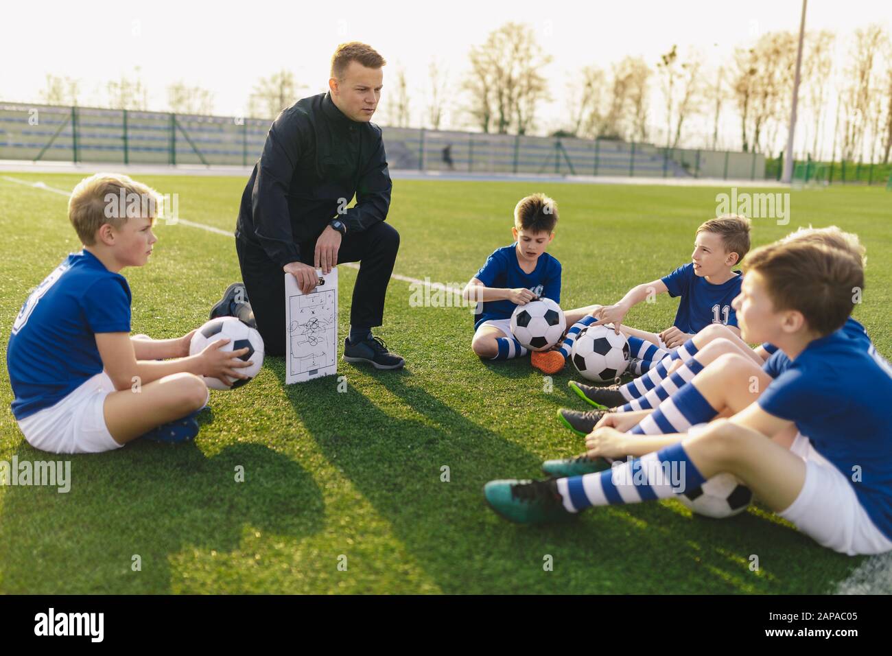 Group of Young Boys Sitting on Sports Grass Field witch School Coach. Kids Listening Coach's Tactic Talk. Young Coach Explain Football Tactic. Coachin Stock Photo